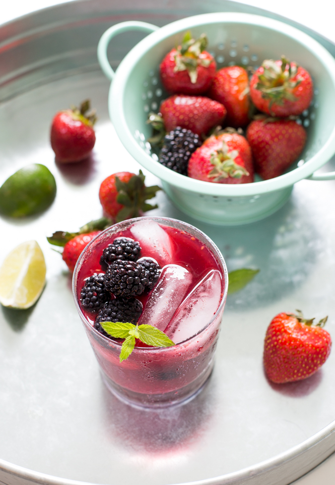 close up of Mixed Berry Mojito garnished with mint, berries and lime scattered on table, colander in background
