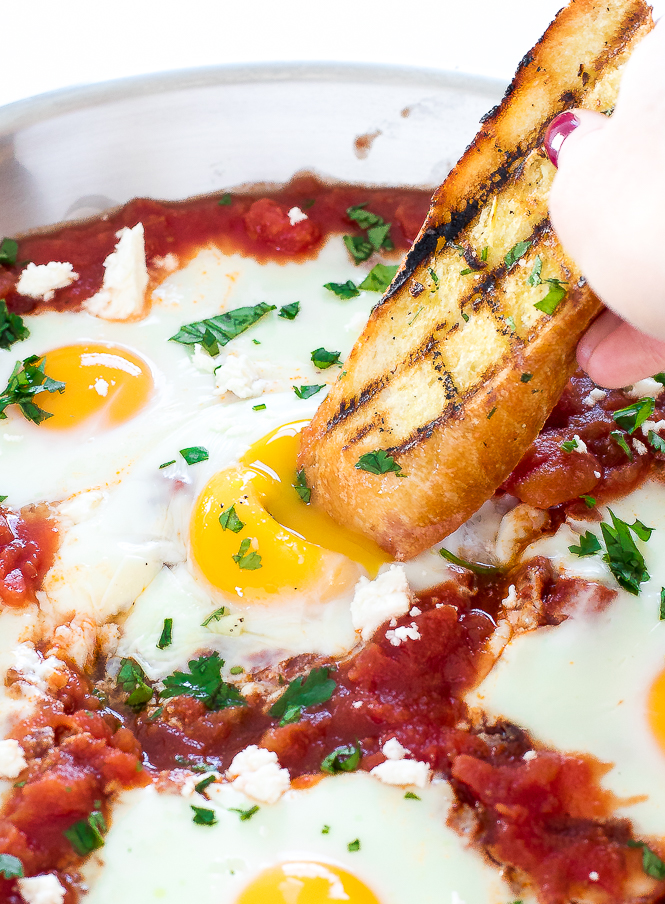 One Skillet Shakshuka author dipping grilled baguette into runny egg yolk of Shakshuka dish