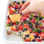 top shot of dip with olives, tomatoes, onions, beans, and sour cream in white baking dish