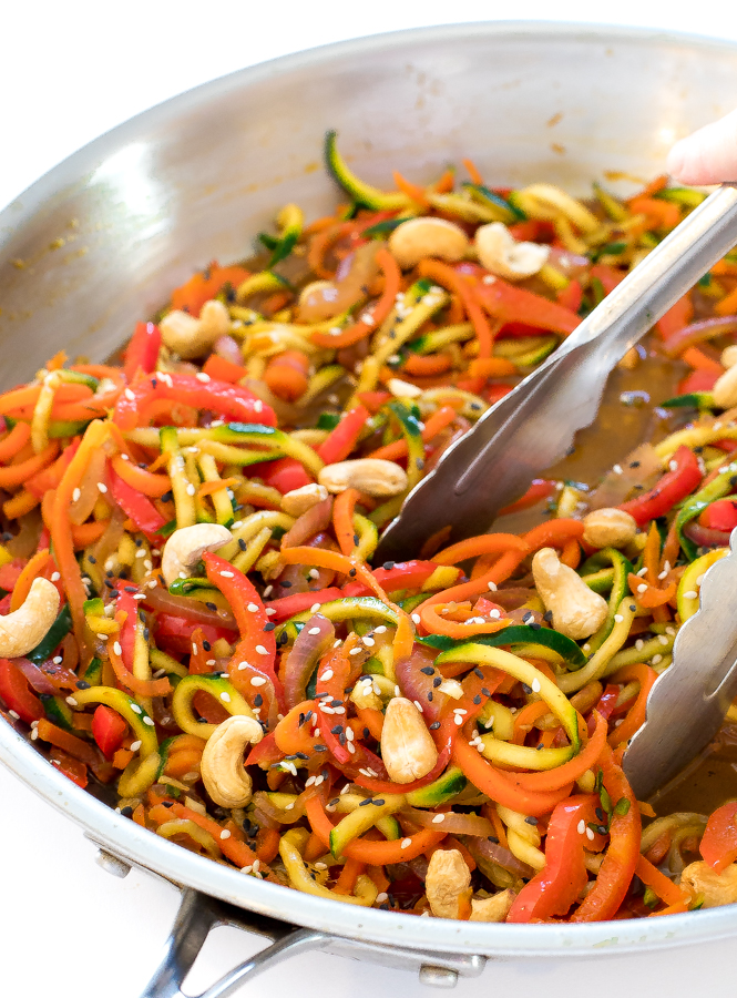 this zucchini noodle stir fry with cashews in a metal skillet with metal tongs tossing the noodles in the sauce 