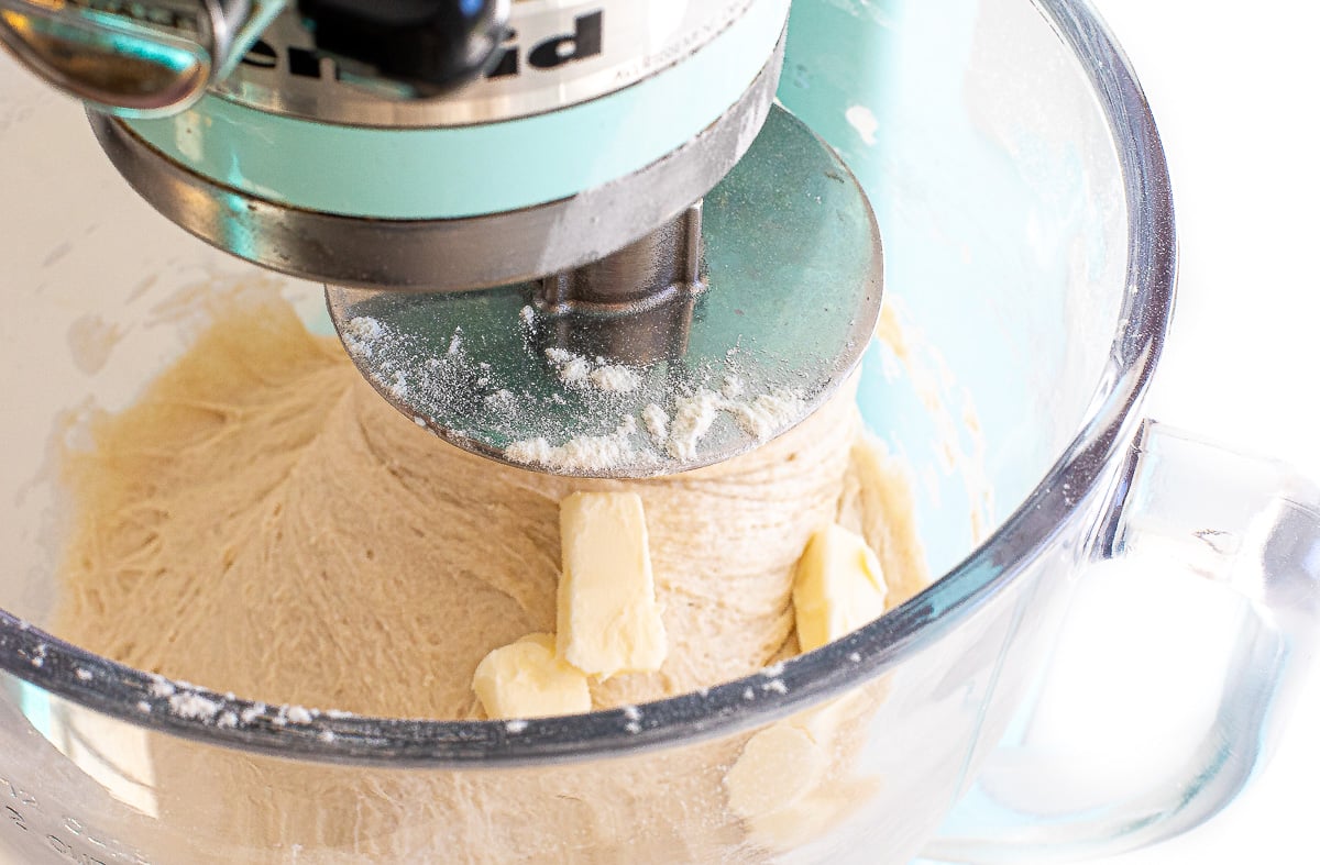 A light blue stand mixer with a dough hook mixing sourdough sandwich bread dough with chunks of butter in a glass bowl.