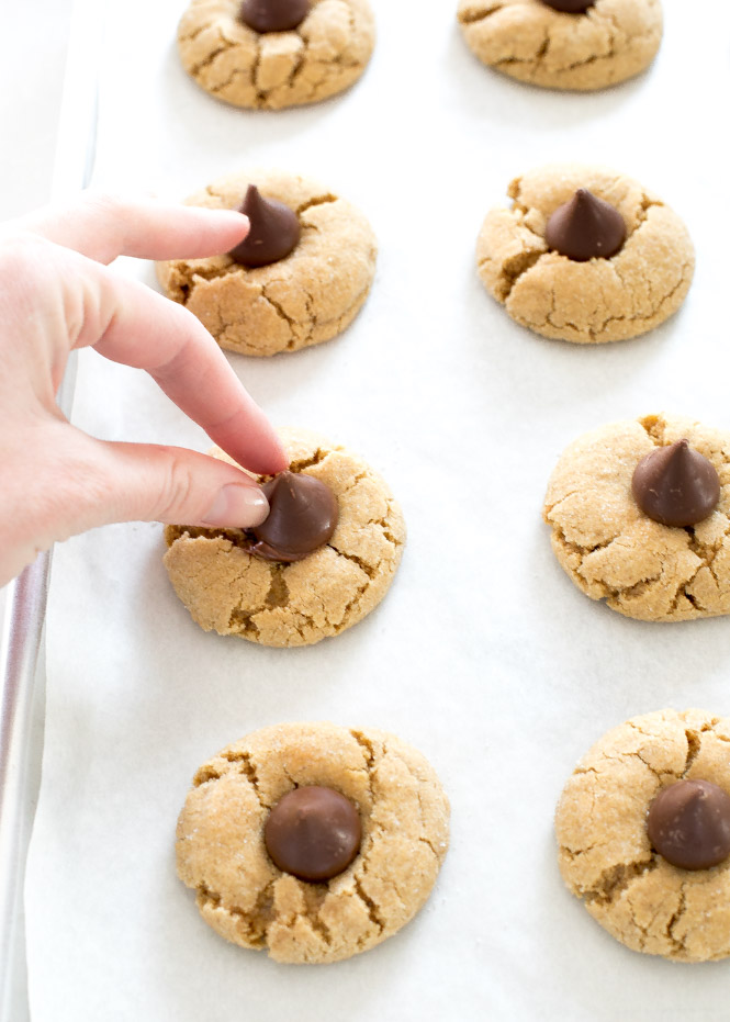 hand placing a Hershey Kiss on a peanut butter blossom cookie surrounded by other cookies on a baking sheet