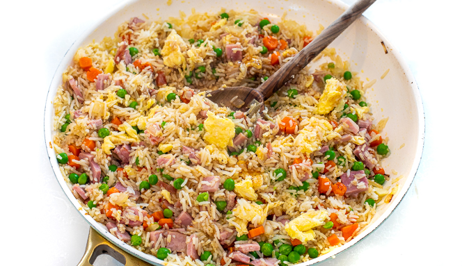 Rice being cooked with veggies and eggs in a white skillet with a wooden spoon.