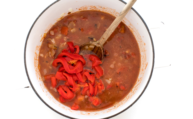 the ingredients for Creamy Roasted Red Pepper and Tomato Soup in a dutch oven with a wooden spoon