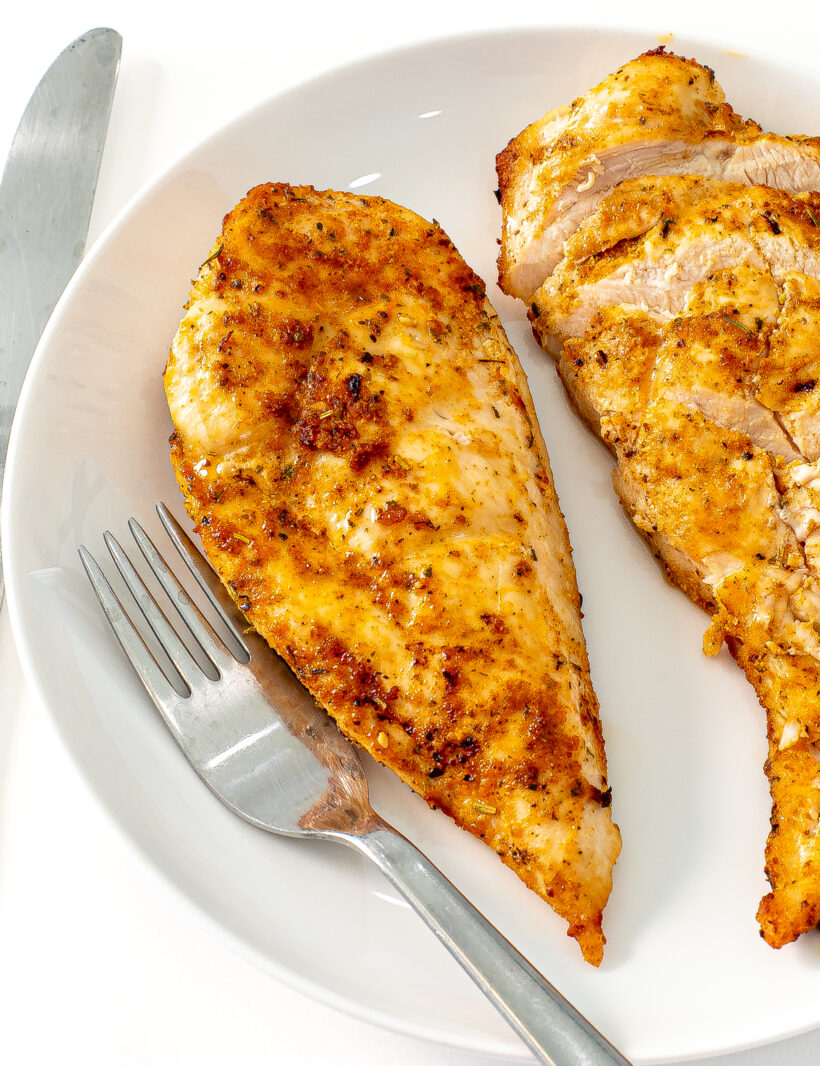 Two chicken breasts on a plate, one is sliced and the other is not, with a fork next to them.