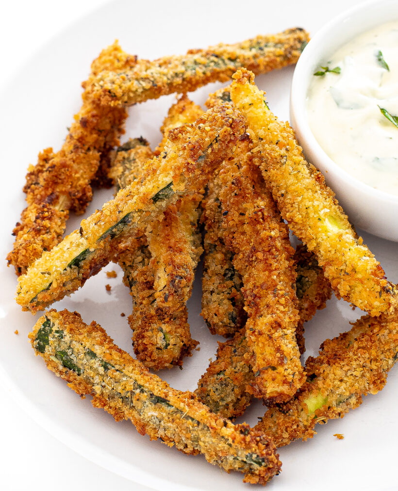 Air fryer zucchini fries on a white plate next to a small bowl of dipping sauce.