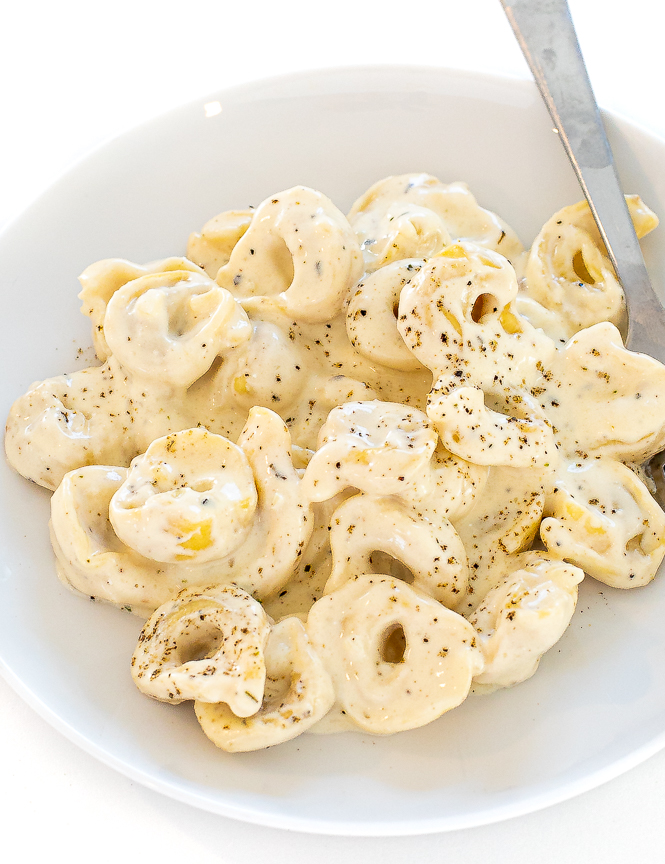 Tortellini Alfredo in a white bowl with pepper on top and silver fork on the side. 