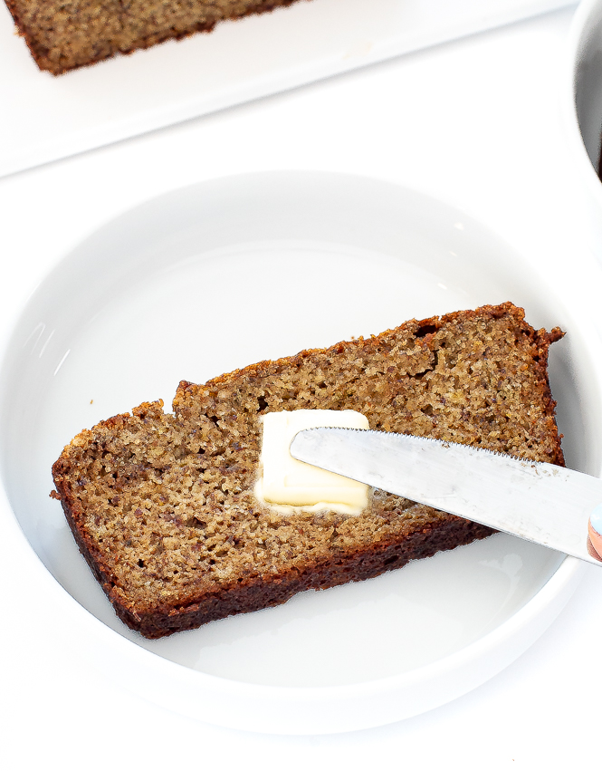 Piece of bread on a plate with butter being spread on top.