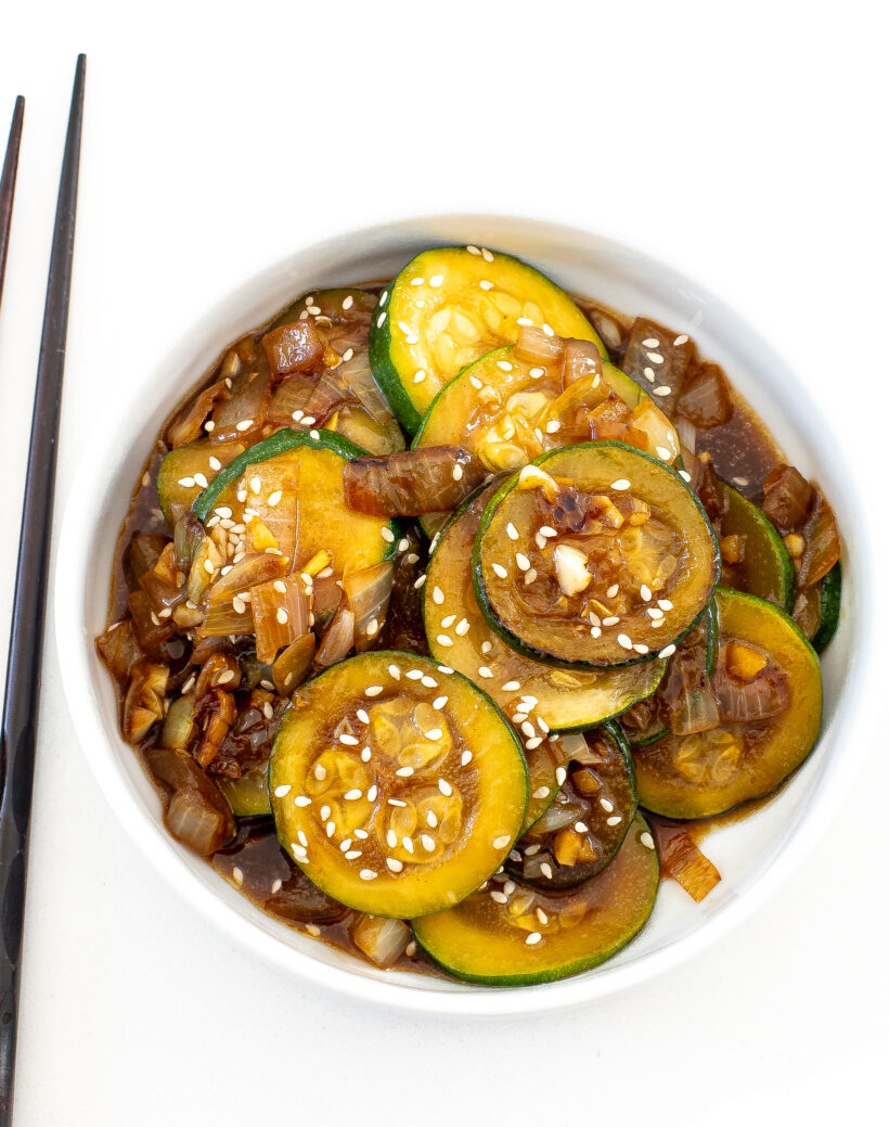 A top down view of a white bowl filled with a serving of zucchini stir fry. Black chop sticks are next to the bowl.