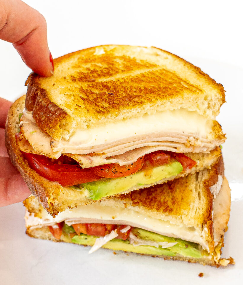 Hand holding a half of a California grilled cheese to show the layer inside with the other half on a plate. 