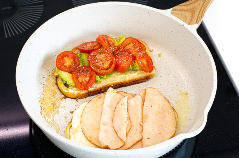 Two slices of bread in a pan, one with avocado and tomato and the other with cheese and turkey.