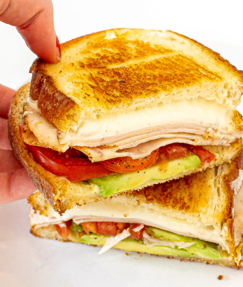 Hand holding half of a California grilled cheese with the other half on a plate. 