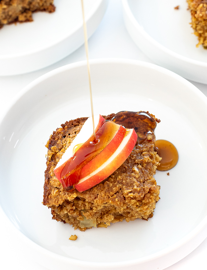 Baked Apple Oatmeal on a white plate with sliced apples, drizzled with maple syrup.