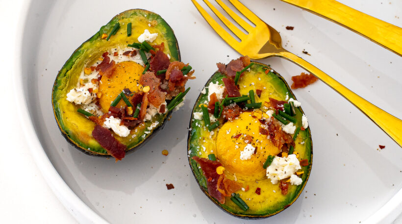 Two avocado halves baked with eggs and topped with bacon and feta on a plate.