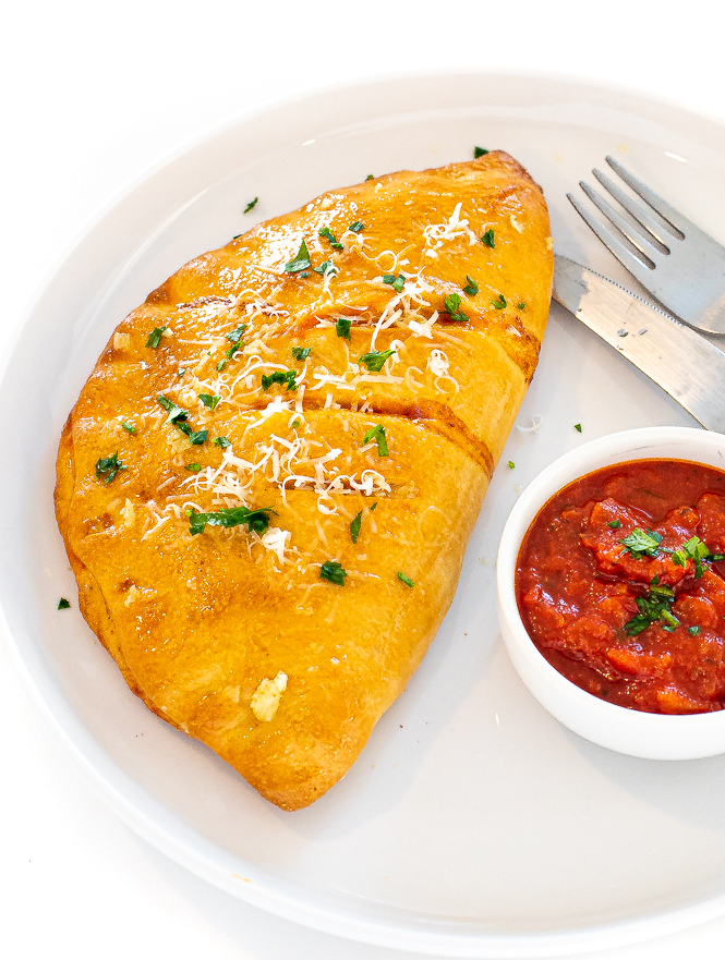 Calzone on a white plate with dipping sauce and fork and knife next to it.