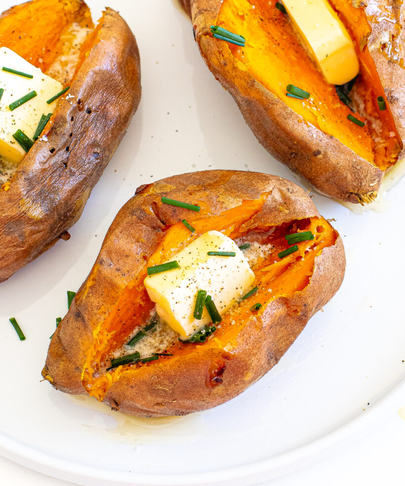 Three sweet potatoes on a white plate topped with butter, chives, and salt and pepper.