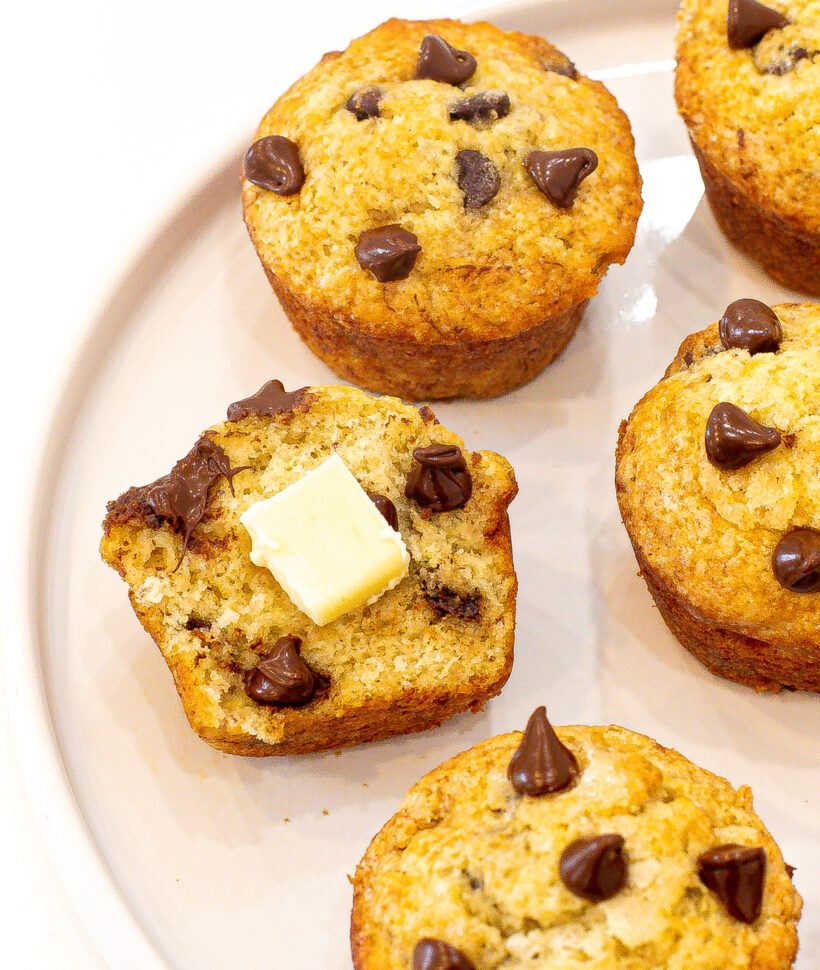 Chocolate chip muffins on a plate with one cut in half and served with a square of butter.