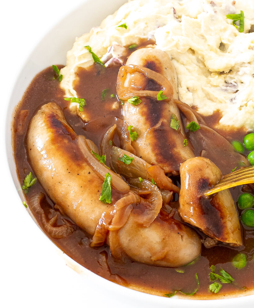Bangers and mash served in a white plate with one of the sausages cut and a fork taking a bite.
