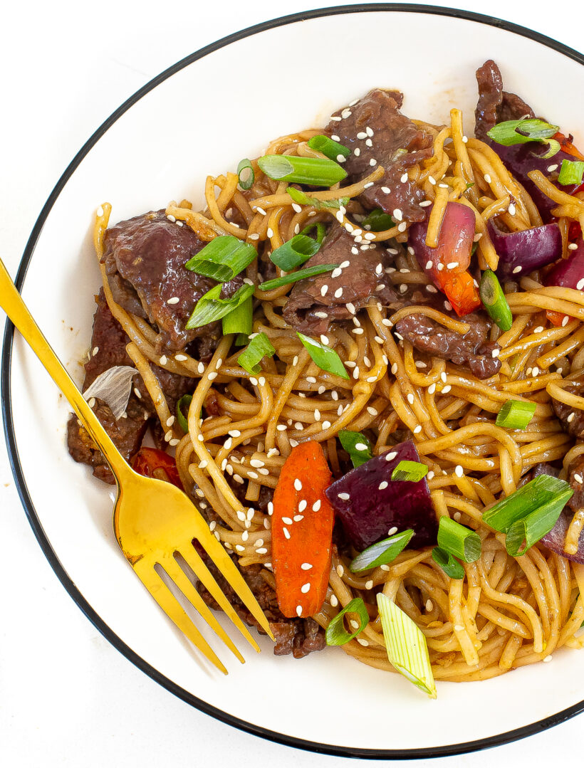 A plate with a serving of beef lo mein topped with sesame seeds and green onions.