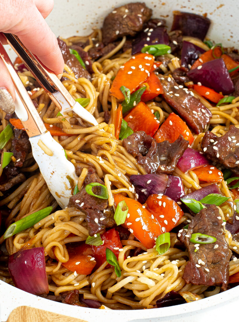 A hand holding tongs and picking up beef lo mein out of the skillet.