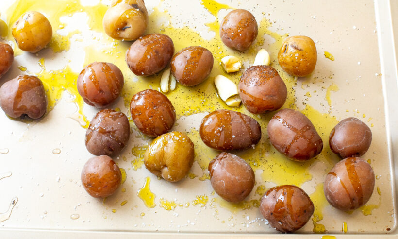 Potatoes on a baking sheet with garlic cloves drizzled with olive oil.
