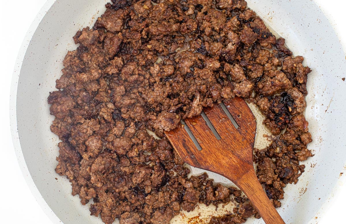 Ground meat browning in a white skillet with a wooden spatula.
