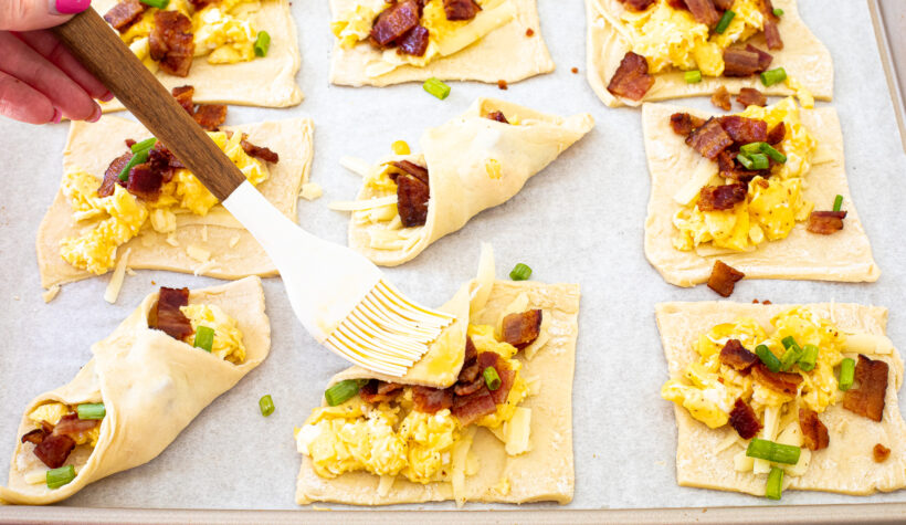 Puff pastry squares with toppings laid out on a baking sheet lined with parchment paper. A hand is brushing the pastries with egg wash.