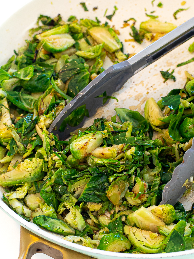 Brussels sprouts chopped up and in a sauté pan with grey tongs. 