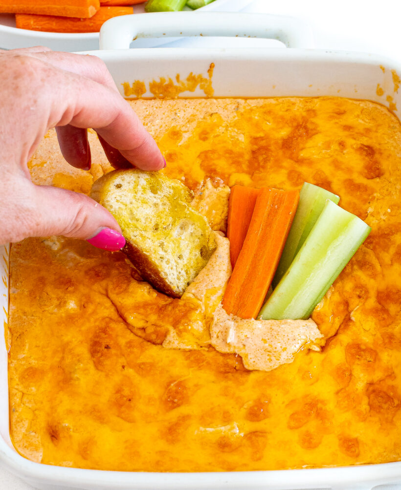 Hand dipping a piece of bread into buffalo chicken dip with carrots and celery also being dipped.