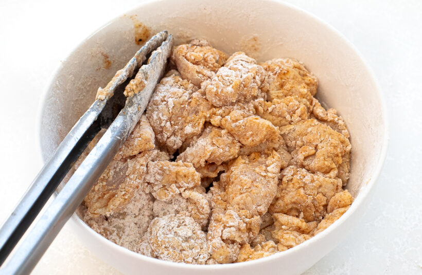 Raw chicken pieces being coated in flour and spices in a mixing bowl.