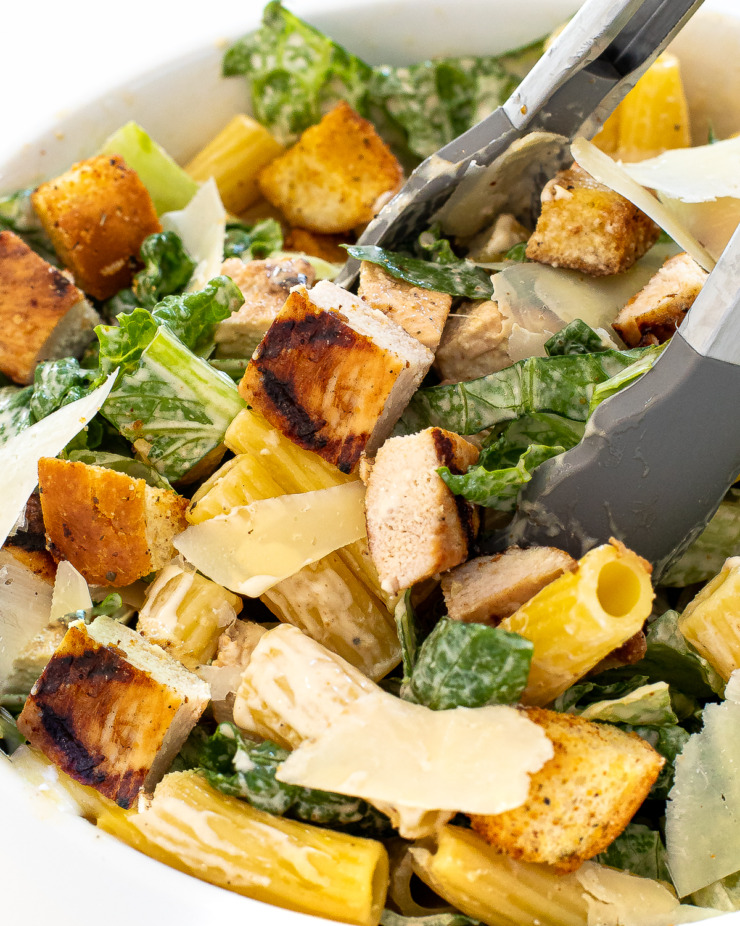 Chicken caesar pasta salad in a large white bowl with tongs.