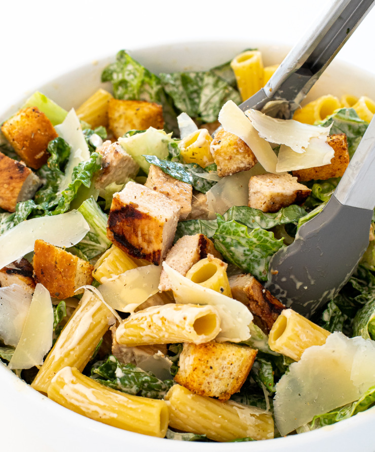 Chicken caesar pasta salad in a white bowl with tongs grabbing a portion.