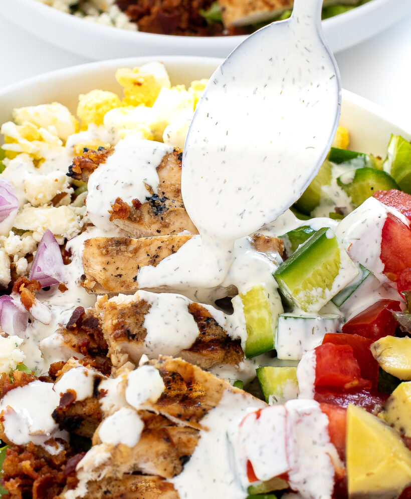 A spoon drizzling ranch dressing over a cobb salad.