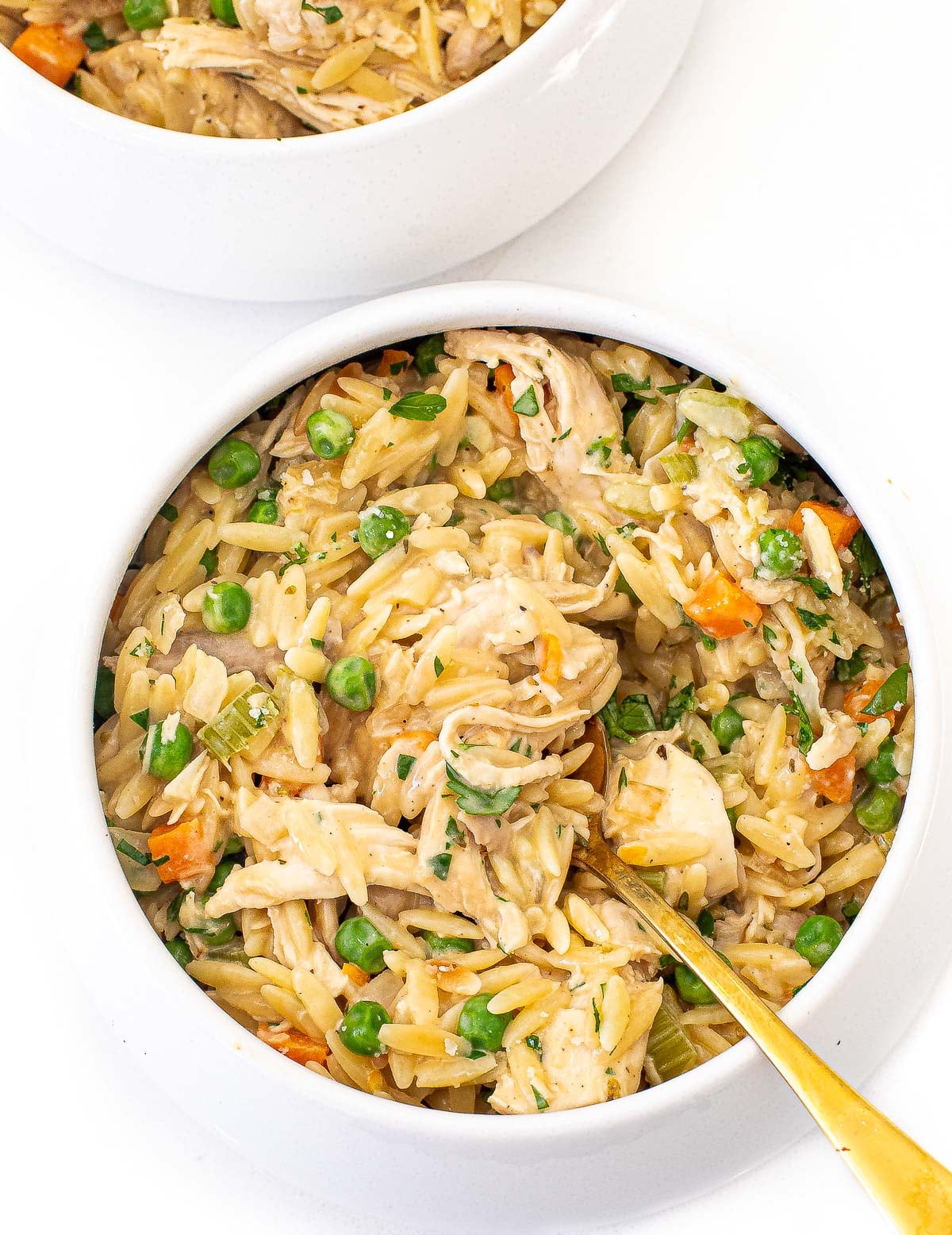 A bowl of chicken orzo soup with peas, carrots, celery, and melting potatoes, with a gold spoon resting inside.