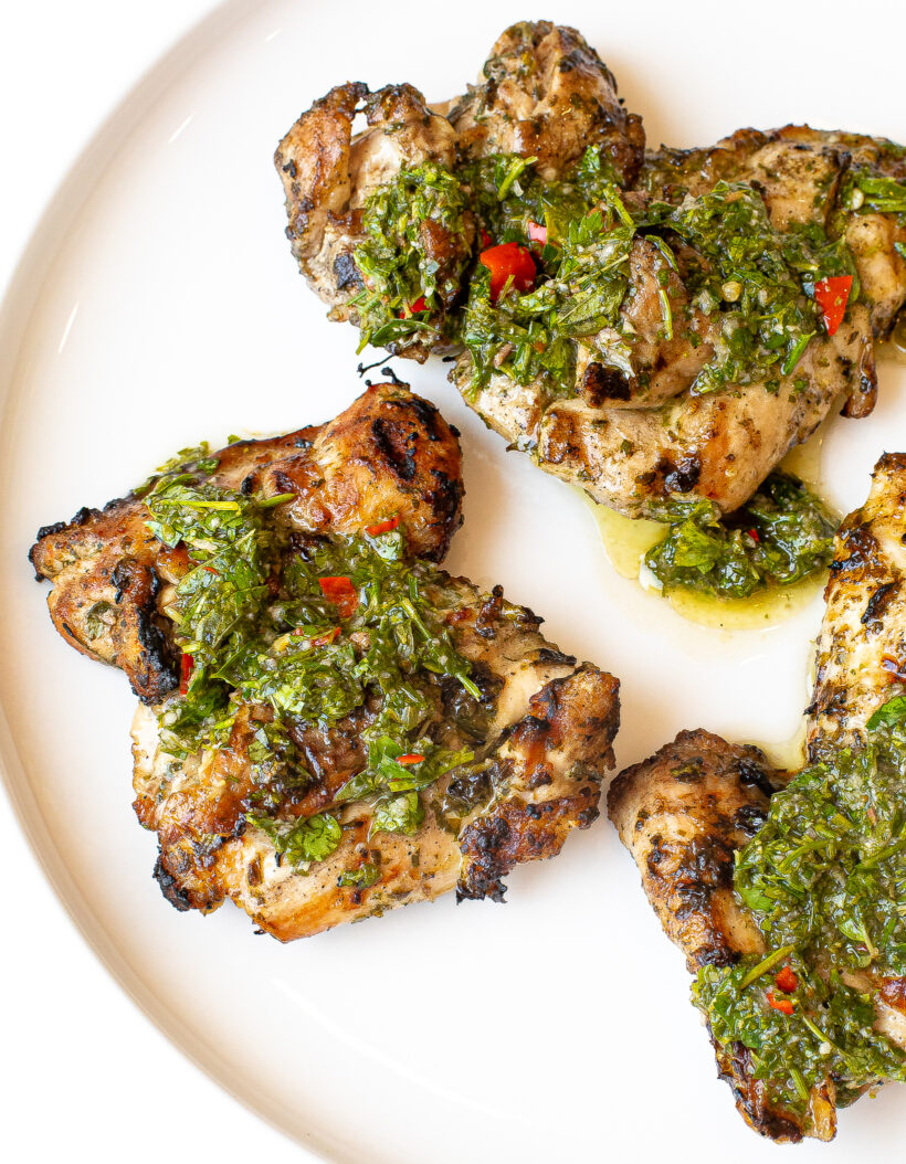 Grilled chicken thighs topped with chimichurri sauce on a white plate.
