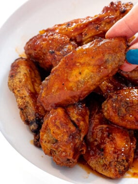 A hand with blue nail polish holds a saucy Honey Chipotle Wing above a white bowl filled with glazed, crispy chicken wings.