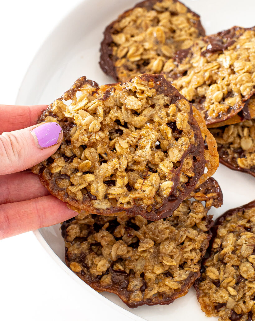 Hand holding a lace cookie sandwich with more cookies in a white bowl. 