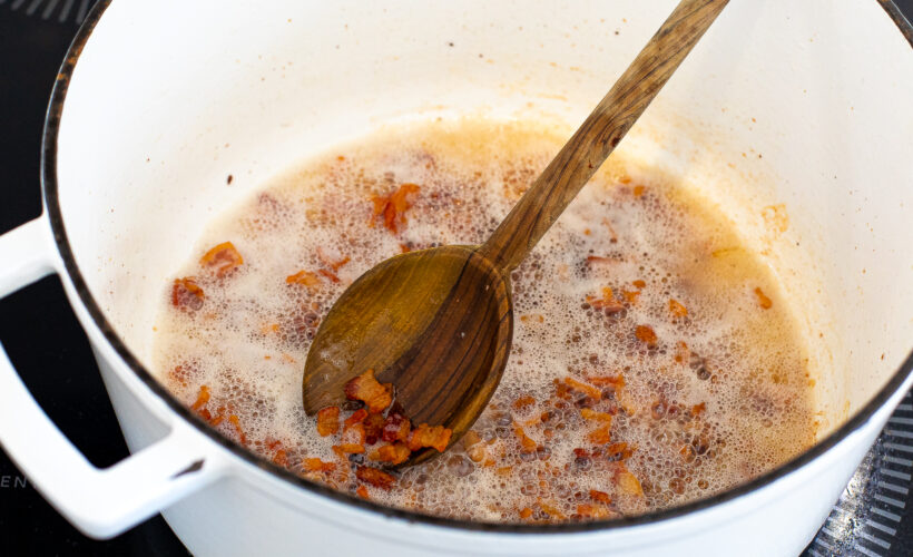 Bacon being sautéed in a dutch oven with a wooden spoon. 