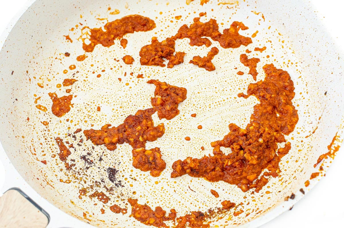 A white non-stick skillet containing clusters of cooked red chili paste and oil.