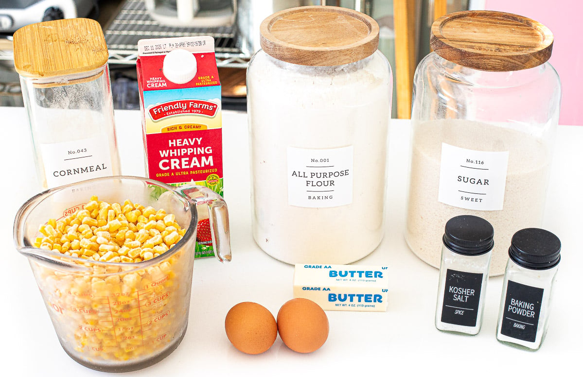 A measuring cup filled with corn kernels sits on a counter next to a carton of whipping cream, two eggs, a stick of butter, and labeled glass jars containing cornmeal, flour, sugar, salt, and baking powder.
