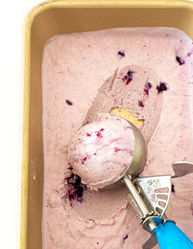 Ice cream scooper scooping blueberry ice cream out of a gold loaf pan. 