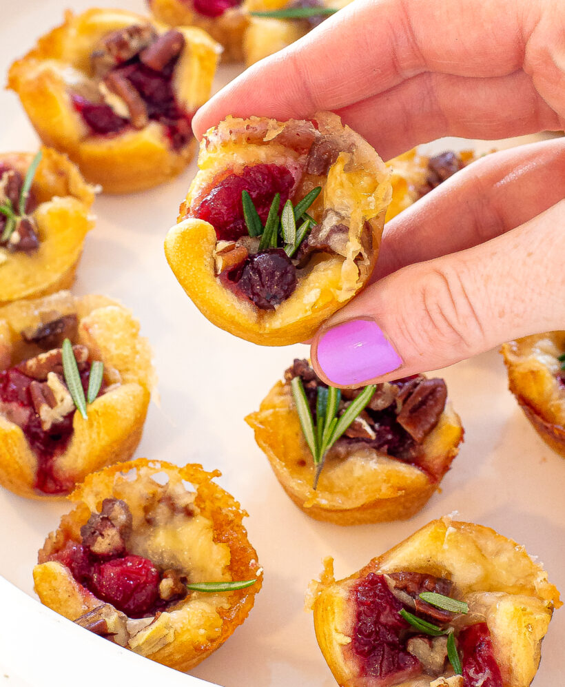 Hand holding one cranberry brie bite topped with fresh rosemary with more in the background. 