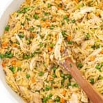 A wooden spoon rests in a skillet of creamy chicken, orzo, peas, carrots, and melting potatoes.