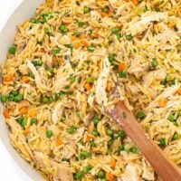 A wooden spoon rests in a skillet of creamy chicken, orzo, peas, carrots, and melting potatoes.