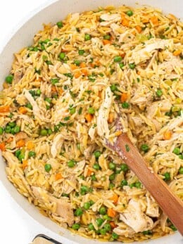 A wooden spoon rests in a skillet of creamy chicken, orzo, peas, carrots, and melting potatoes.