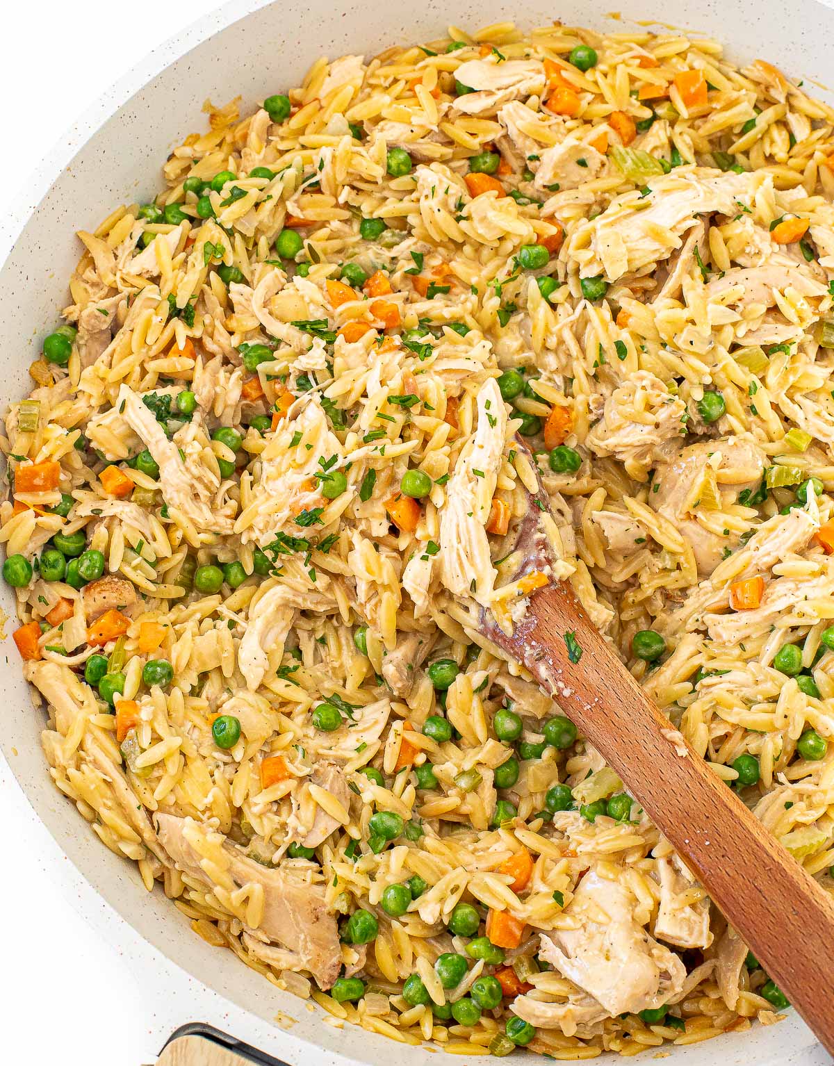 A wooden spoon rests in a skillet of creamy chicken, orzo, peas, carrots, and melting potatoes.