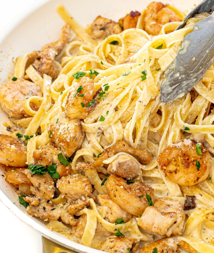 Silver tongs taking fettuccini alfredo from a skillet.