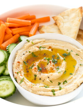 Small bowl of hummus with oil on top on platter with pita and veggies.