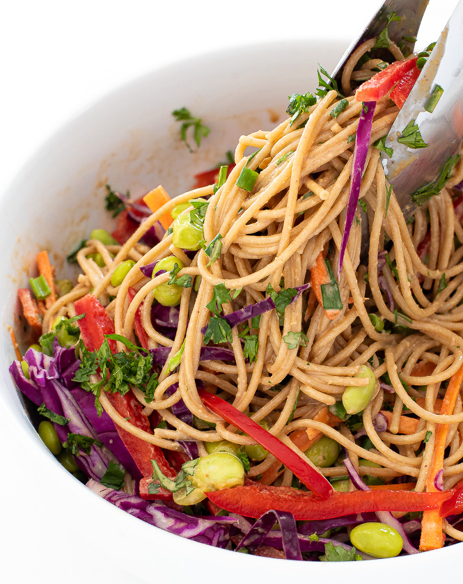 Tongs grabbing the rainbow peanut noodles from a white serving bowl.