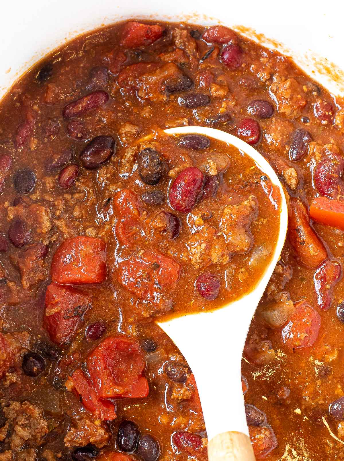 A close-up of Slow Cooker Chili in a pot, showing beans, ground meat, and tomato chunks in a rich, red sauce. A white spoon is partially submerged, scooping up the chili.